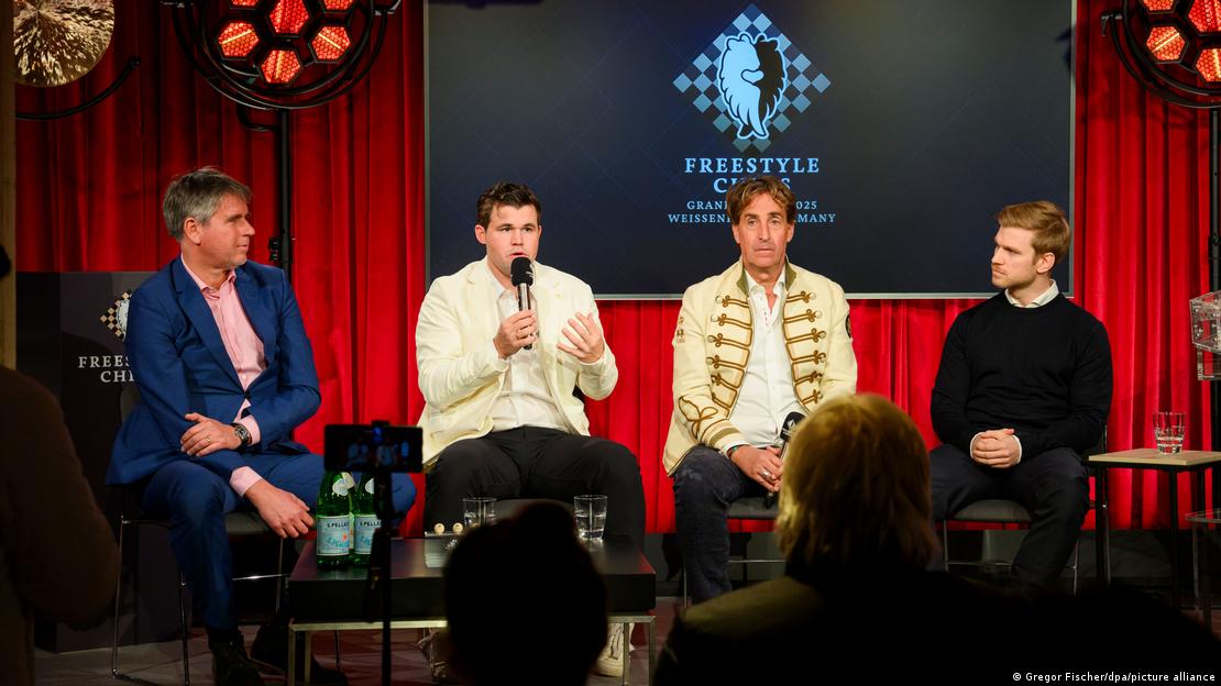 Four men on a stage in front of red curtains and a Freestyle Chess Grand Slam backdrop; Magnus Carlsen, in a white jacket, speaks into a microphone.