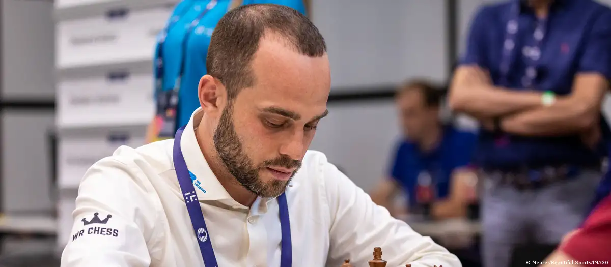A chess player in a white shirt and tournament lanyard concentrates on the board.
