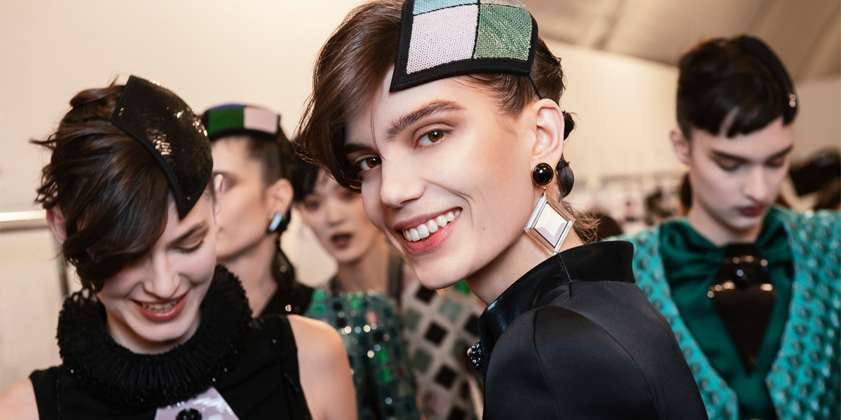 Backstage, four smiling models with sequinned geometric headpieces wear black and green Armani looks.