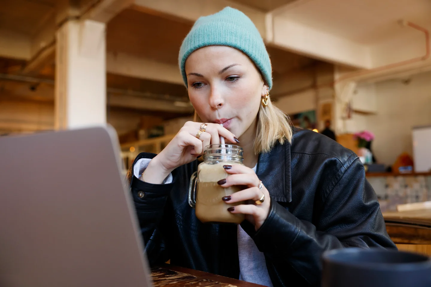 A young woman in a teal beanie sips an iced coffee through a straw while looking at a laptop in a café.